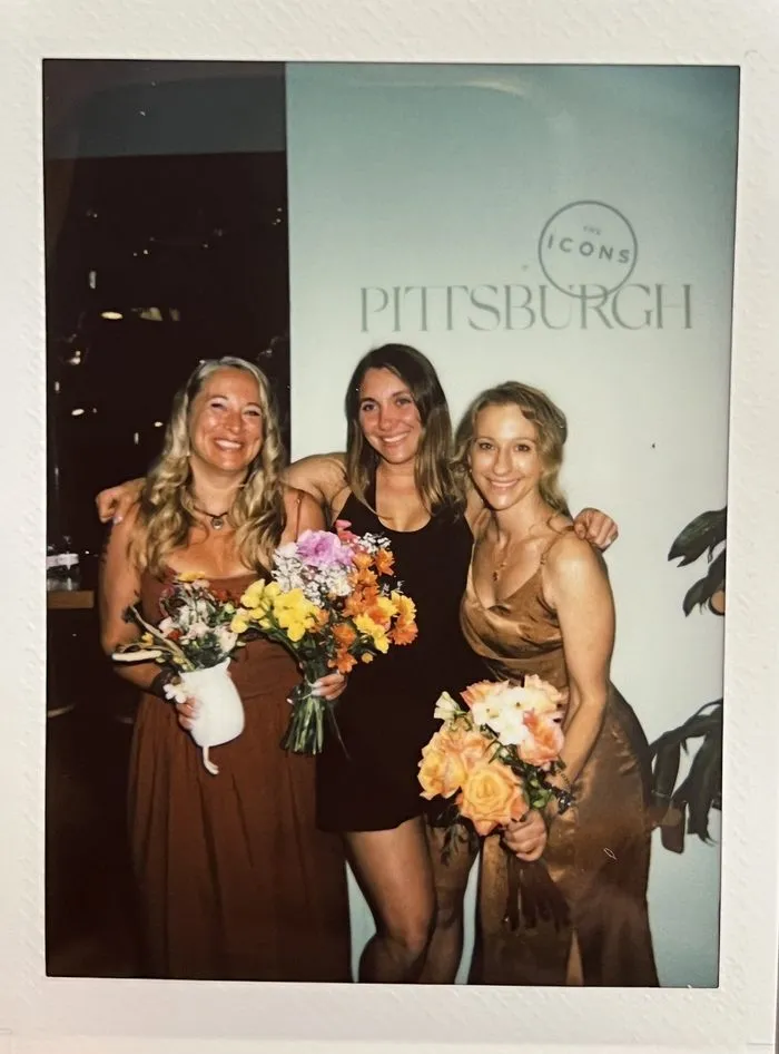 Three women holding flowers in front of The ICONS of Pittsburgh step-and-repeat banner